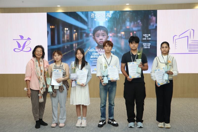Five young people holding certificates and small bouquets stand in a row beside a woman in a pink jacket, in front of a large screen that reads Nurture Our Future.