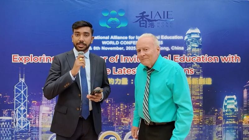 Two men stand in front of a conference backdrop showing the Hong Kong skyline: one, in a suit, holds a microphone, and the other wears a turquoise shirt and a striped tie.