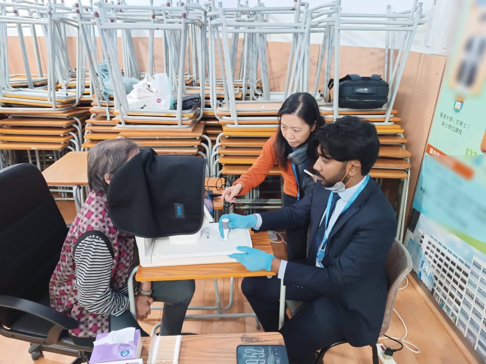 A volunteer in a suit and blue gloves works the joystick of a fundus camera while a visitor looks into it under a dark cloth and a second helper leans in to point at the controls.
