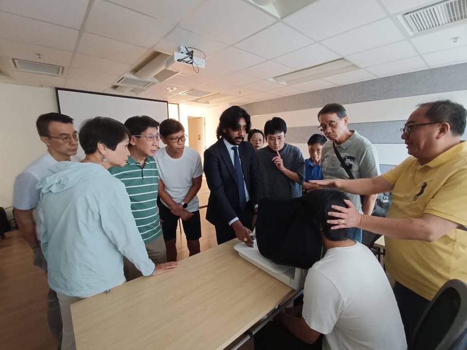 A group leans in around a table as a man in a suit points out the controls of a fundus camera and another man steadies the head of the person seated at it.