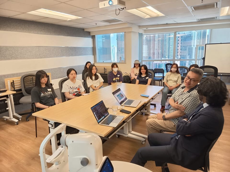 Volunteers sit along the far side of a meeting table with two open laptops, listening to a man speaking at the right, with part of a fundus camera in the foreground.